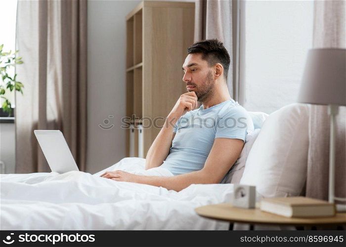 people, technology and remote job concept - man with laptop computer working in bed at home bedroom. man with laptop in bed at home bedroom