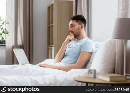 people, technology and remote job concept - man with laptop computer working in bed at home bedroom. man with laptop in bed at home bedroom