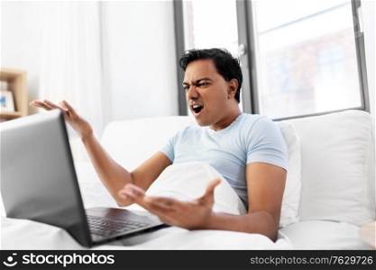 people, technology and rest concept - angry indian man with laptop computer lying in bed at home. angry indian man with laptop in bed at home