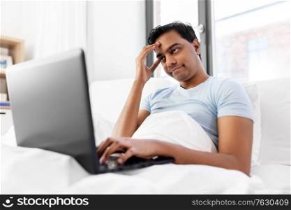 people, technology and rest concept - bored indian man with laptop computer lying in bed at home. bored indian man with laptop in bed at home