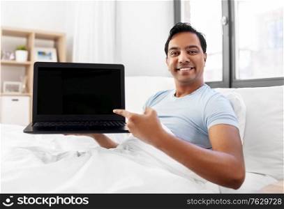 people, technology and rest concept - happy smiling indian man showing his laptop computer in bed at home. happy indian man showing his laptop in bed at home