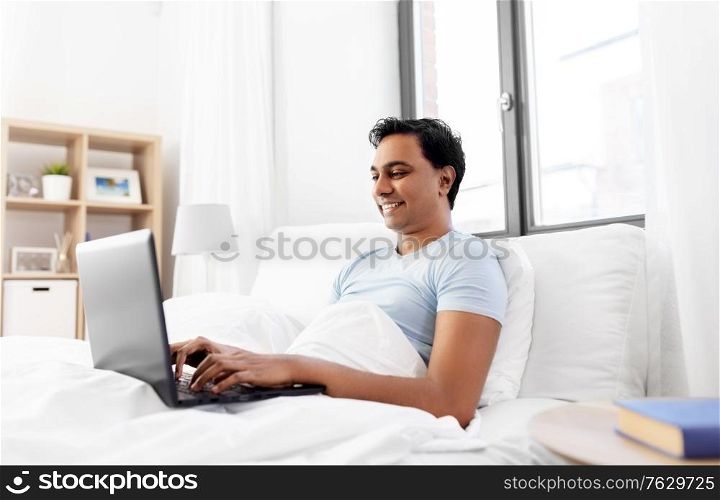 people, technology and rest concept - happy smiling indian man with laptop computer lying in bed at home. happy indian man with laptop in bed at home