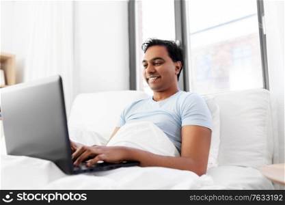 people, technology and rest concept - happy smiling indian man with laptop computer lying in bed at home. happy indian man with laptop in bed at home
