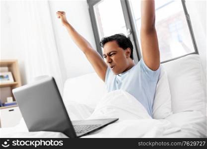 people, technology and success concept - happy smiling indian man with laptop computer lying in bed and making winning gesture at home. happy indian man with laptop in bed at home