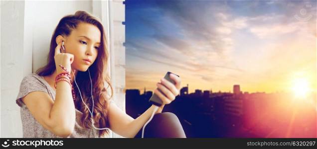 people, technology and teens concept - pretty teenage girl sitting on windowsill with smartphone and earphones listening to music over city background. teenage girl with smartphone and earphones