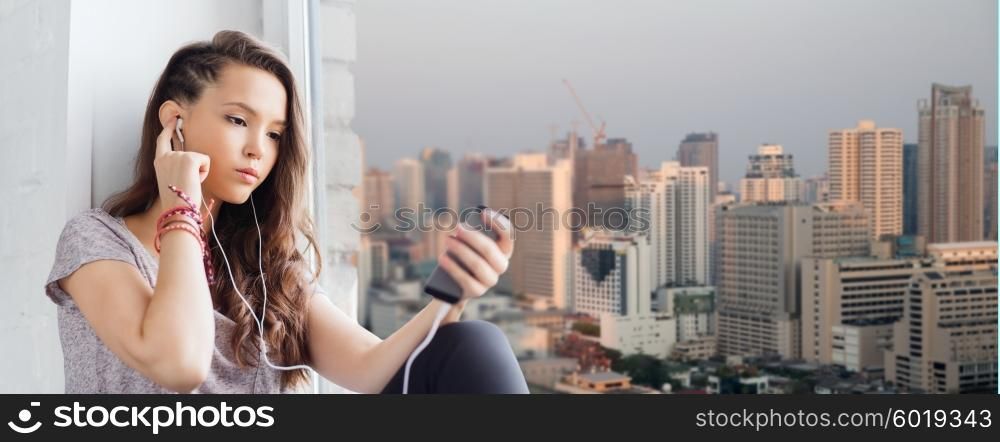 people, technology and teens concept - sad pretty teenage girl sitting on windowsill with smartphone and earphones listening to music over city background