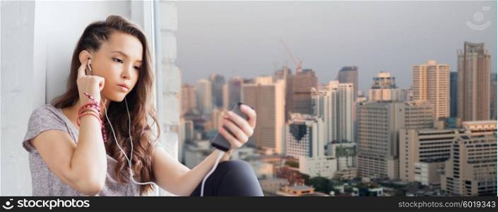 people, technology and teens concept - sad pretty teenage girl sitting on windowsill with smartphone and earphones listening to music over city background