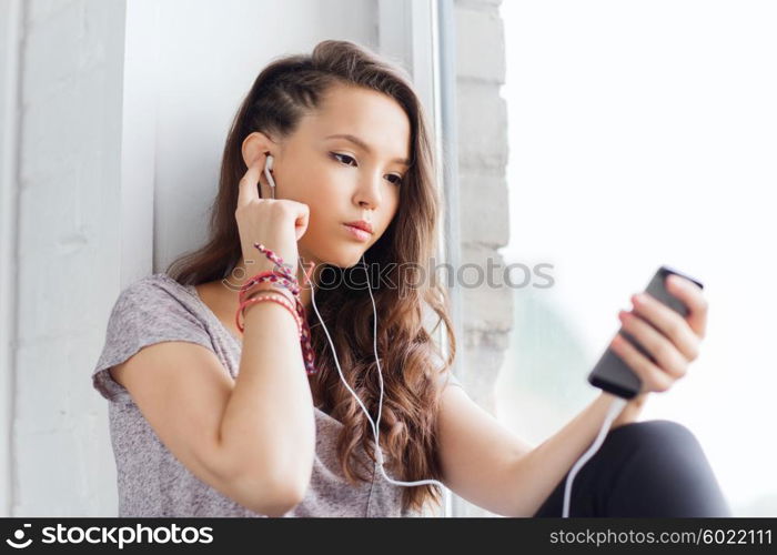 people, technology and teens concept - sad pretty teenage girl sitting on windowsill with smartphone and earphones listening to music