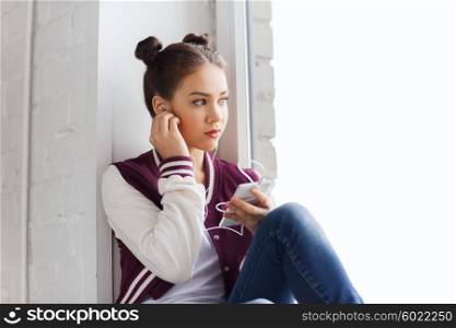 people, technology and teens concept - sad pretty teenage girl sitting on windowsill with smartphone and earphones listening to music