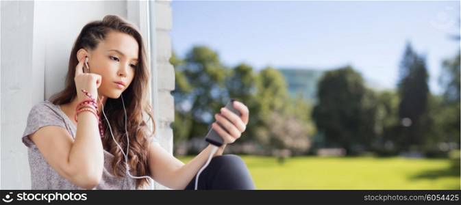 people, technology and teens concept - sad pretty teenage girl sitting on windowsill with smartphone and earphones listening to music over summer background