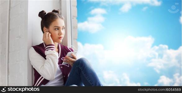 people, technology and teens concept - sad pretty teenage girl sitting on windowsill with smartphone and earphones listening to music over blue sky and clouds background