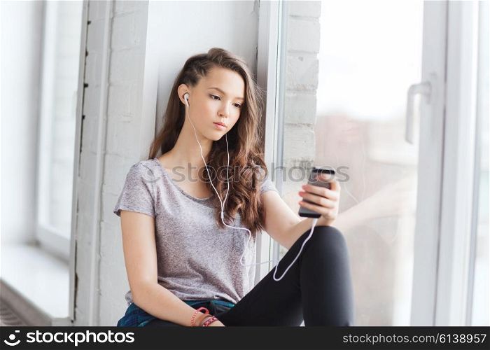 people, technology and teens concept - sad pretty teenage girl sitting on windowsill with smartphone and earphones listening to music
