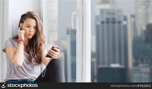people, technology and teens concept - sad pretty teenage girl sitting on windowsill with smartphone and earphones listening to music over singapore city skyscrapers background