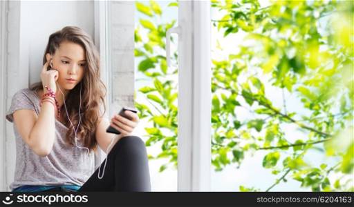 people, technology and teens concept - sad pretty teenage girl sitting on windowsill with smartphone and earphones listening to music over green natural background