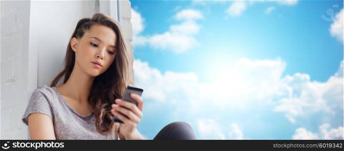 people, technology and teens concept - sad unhappy pretty teenage girl sitting on windowsill with smartphone and texting over blue sky and clouds background