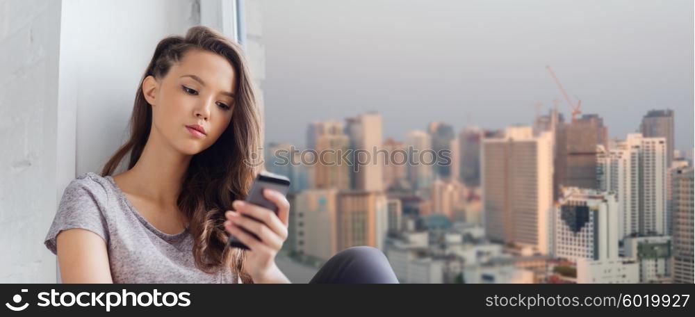 people, technology and teens concept - sad unhappy pretty teenage girl sitting on windowsill with smartphone and texting over city background