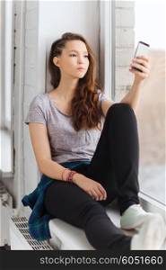 people, technology and teens concept - sad unhappy pretty teenage girl sitting on windowsill with smartphone and texting
