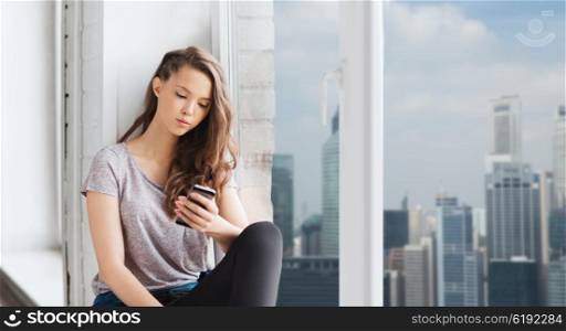 people, technology and teens concept - sad unhappy pretty teenage girl sitting on windowsill with smartphone and texting over singapore city skyscrapers background