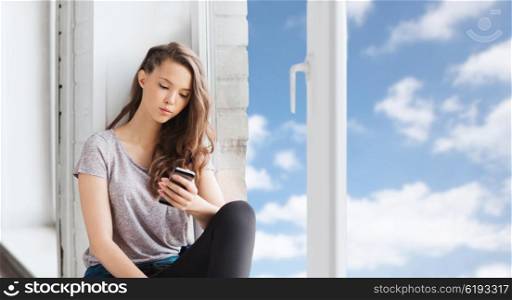 people, technology and teens concept - sad unhappy pretty teenage girl sitting on windowsill with smartphone and texting over blue sky and clouds background