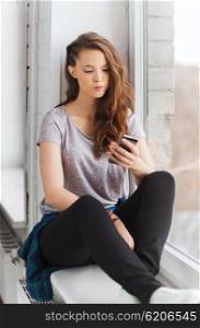 people, technology and teens concept - sad unhappy pretty teenage girl sitting on windowsill with smartphone and texting