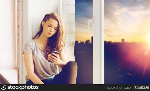people, technology and teens concept - sad unhappy pretty teenage girl sitting on windowsill with smartphone and messaging over city background. sad pretty teenage girl with smartphone texting