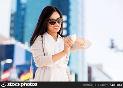 people, technology and time management concept - smiling young asian woman in sunglasses looking at her wristwatch or smart watch in city. woman looking at wristwatch or smart watch in city