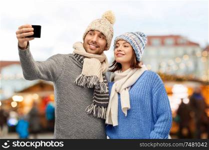 people, technology and winter holidays concept - happy couple in knitted hats and scarves taking selfie by smartphone over christmas market in old town of tallinn city background. couple takes selfie by smartphone christmas market