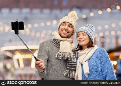 people, technology and winter holidays concept - happy couple in knitted hats and scarves taking picture by smartphone on selfie stick over christmas market and lights background. happy couple in winter clothes taking selfie