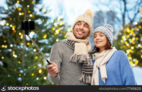 people, technology and winter holidays concept - happy couple in knitted hats and scarves taking picture by smartphone on selfie stick outdoors over christmas tree lights background. happy couple in winter clothes taking selfie