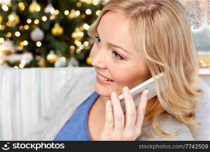 people, technology and winter holidays concept - smiling middle aged woman calling on smartphone at home over christmas tree lights on background. smiling woman calling on smartphone on christmas