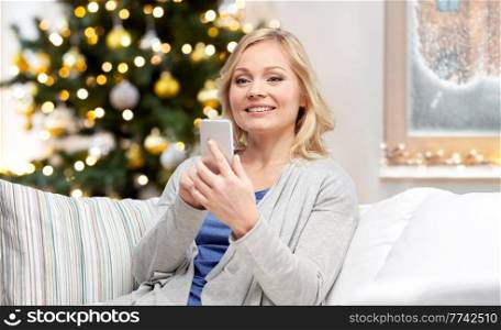 people, technology and winter holidays concept - smiling middle aged woman with smartphone texting at home over christmas tree lights on background. smiling woman with smartphone at home on christmas
