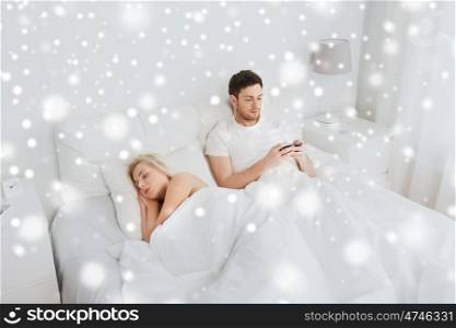 people, technology, cheating, internet addiction and communication concept - man with smartphone texting message while woman is sleeping in bed over snow