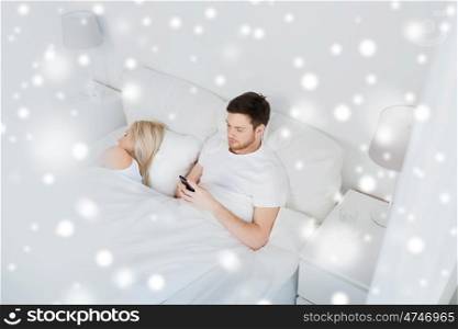 people, technology, cheating, internet addiction and communication concept - man with smartphone texting message while woman is sleeping in bed over snow