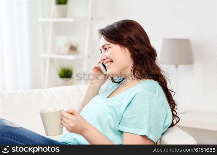 people, technology, communication and leisure concept - happy plus size young woman with cup of coffe or tea calling on smartphone at home