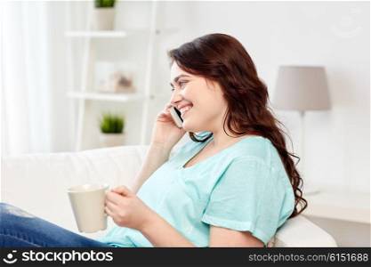 people, technology, communication and leisure concept - happy plus size young woman with cup of coffe or tea calling on smartphone at home