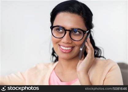people, technology, communication and leisure concept - happy smiling young indian woman in glasses calling on smartphone at home. happy indian woman calling on smartphone at home