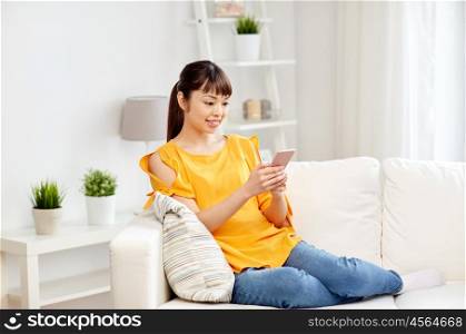 people, technology, communication and leisure concept - happy young asian woman sitting on sofa and texting message on smartphone at home