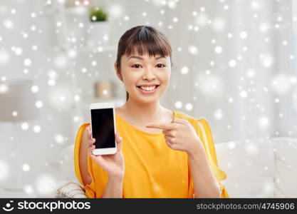 people, technology, communication and leisure concept - happy young asian woman sitting on sofa and showing smartphone blank screen at home over snow