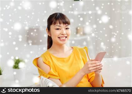 people, technology, communication and leisure concept - happy young asian woman sitting on sofa and texting message on smartphone at home over snow