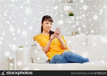 people, technology, communication and leisure concept - happy young asian woman sitting on sofa and texting message on smartphone at home over snow