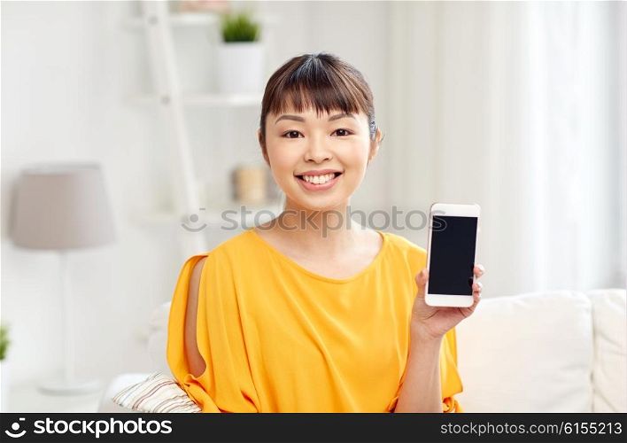people, technology, communication and leisure concept - happy young asian woman sitting on sofa and showing smartphone blank screen at home