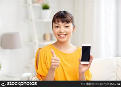 people, technology, communication and leisure concept - happy young asian woman sitting on sofa and showing smartphone blank screen at home