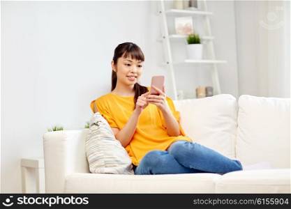 people, technology, communication and leisure concept - happy young asian woman sitting on sofa and texting message on smartphone at home