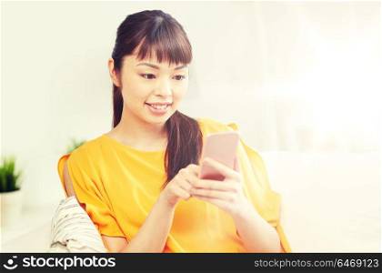people, technology, communication and leisure concept - happy young asian woman sitting on sofa and texting message on smartphone at home. happy asian woman with smartphone at home