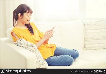 people, technology, communication and leisure concept - happy young asian woman sitting on sofa and texting message on smartphone at home. happy asian woman with smartphone at home. happy asian woman with smartphone at home