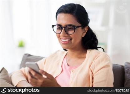 people, technology, communication and leisure concept - happy young indian woman in glasses sitting on sofa and texting message on smartphone at home. happy woman texting message on smartphone at home
