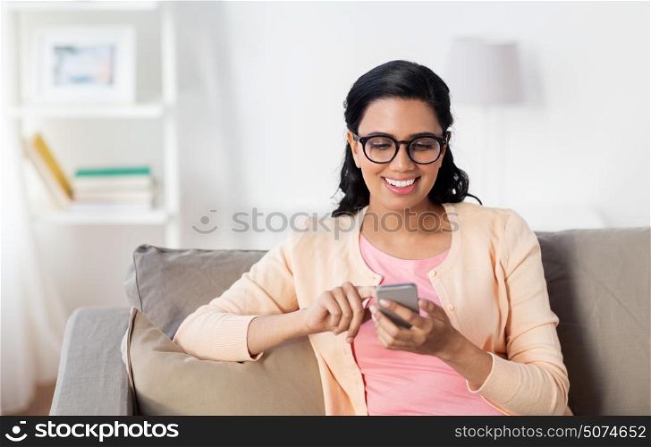 people, technology, communication and leisure concept - happy young indian woman sitting on sofa and texting message on smartphone at home. happy woman texting message on smartphone at home