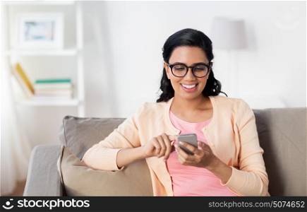 people, technology, communication and leisure concept - happy young indian woman sitting on sofa and texting message on smartphone at home. happy woman texting message on smartphone at home