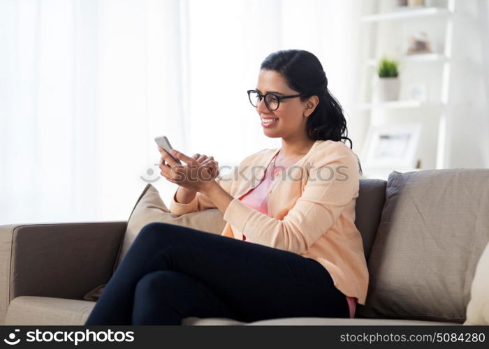 people, technology, communication and leisure concept - happy young indian woman sitting on sofa and texting message on smartphone at home. happy woman texting message on smartphone at home. happy woman texting message on smartphone at home