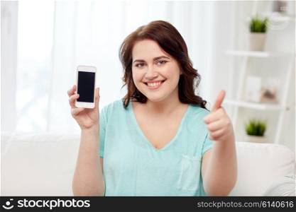 people, technology, communication and leisure concept - happy young plus size woman sitting on sofa and showing smartphone blank screen and showing thumbs up at home
